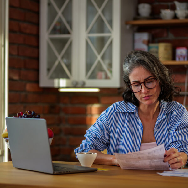 Mid adult woman at home analyzing bills, using her laptop in the kitchen, carefully managing personal finances and budget, experiencing financial stress from remote work